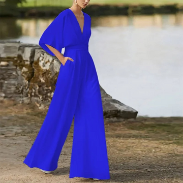 Frau in leuchtend blauem Jumpsuit mit weitem Bein und tiefem V-Ausschnitt, steht draußen vor einem steinernen Hintergrund. Elegante Damenmode.