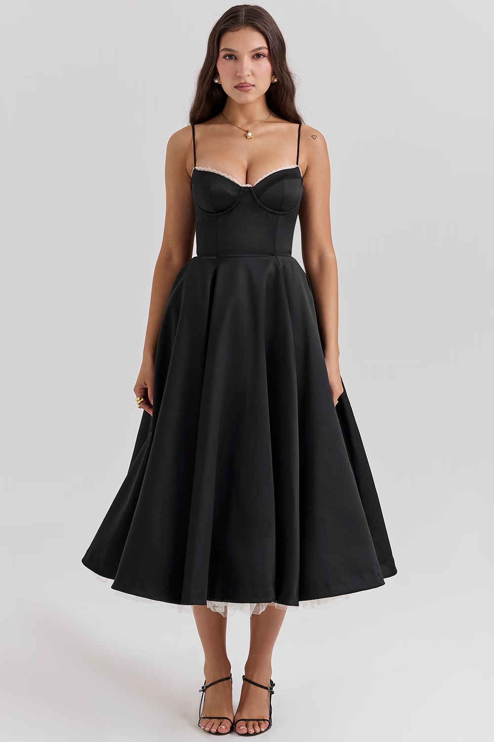 Frau in elegantem, schwarzem Cocktailkleid mit Spaghettiträgern und ausgestelltem Rock, stehend vor grauem Hintergrund. Abendmode, Damenkleidung.