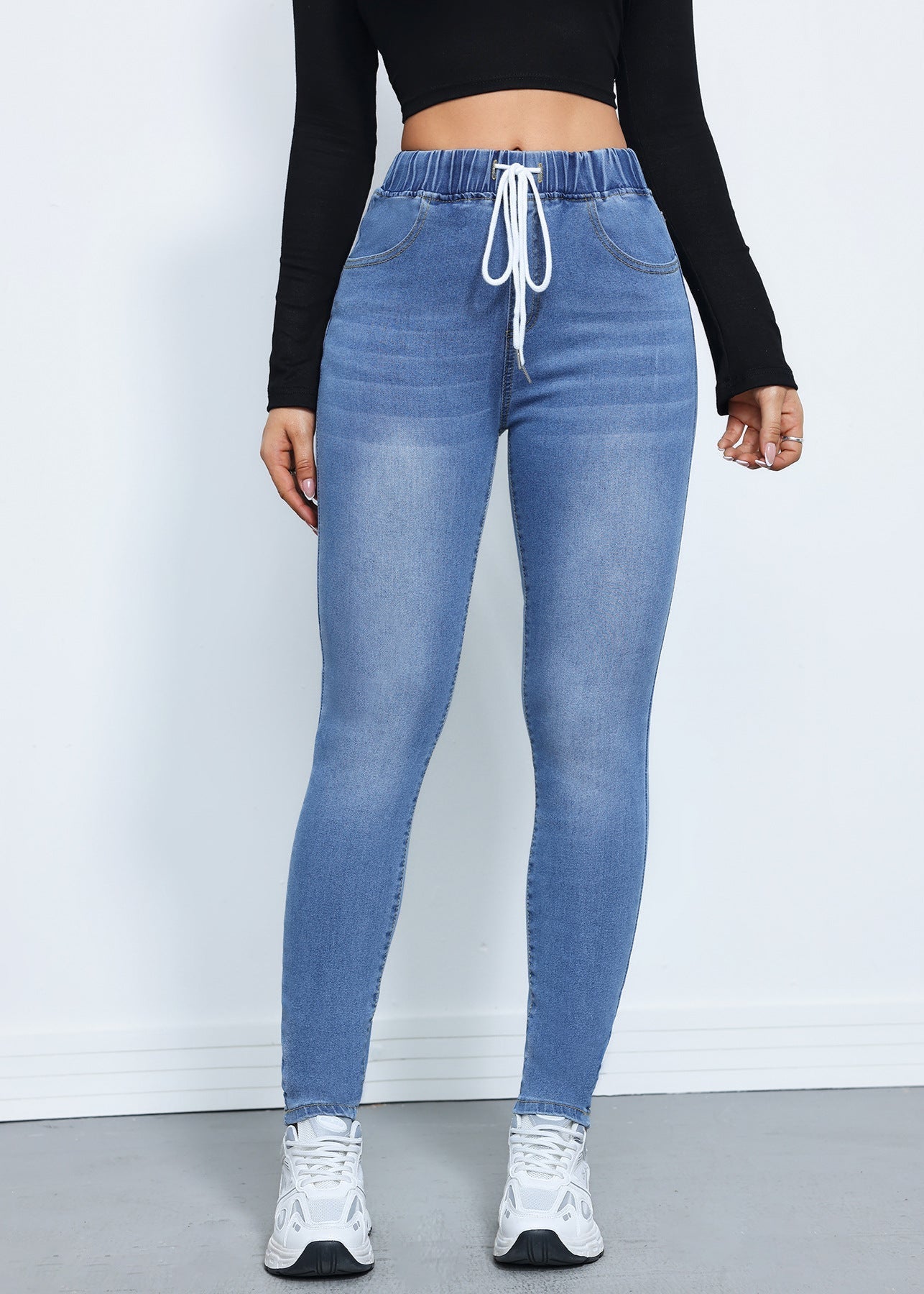 Frau trägt blaue Jeans-Leggings mit Kordelzug, kombiniert mit schwarzem Langarm-Oberteil und weißen Sneakers. Modetrend, Damenbekleidung, Freizeitlook.