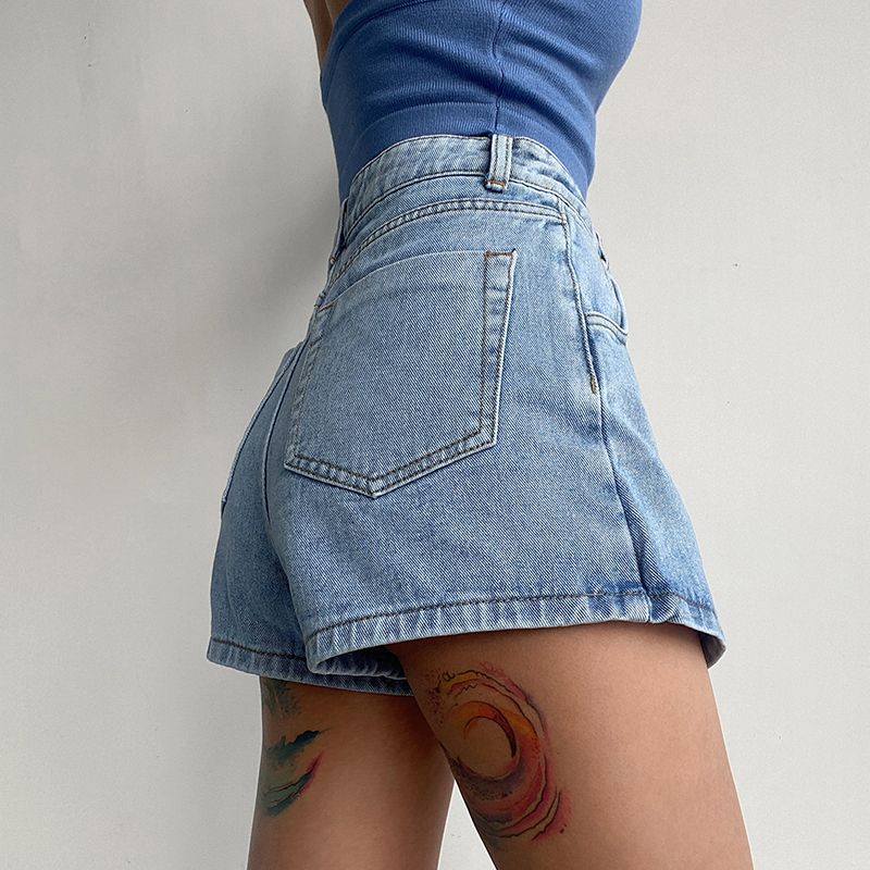 Frau in blauen Jeansshorts und blauem Top, zeigt bunte Tattoos am Oberschenkel. Modetrend, Sommeroutfit, lässiger Stil.