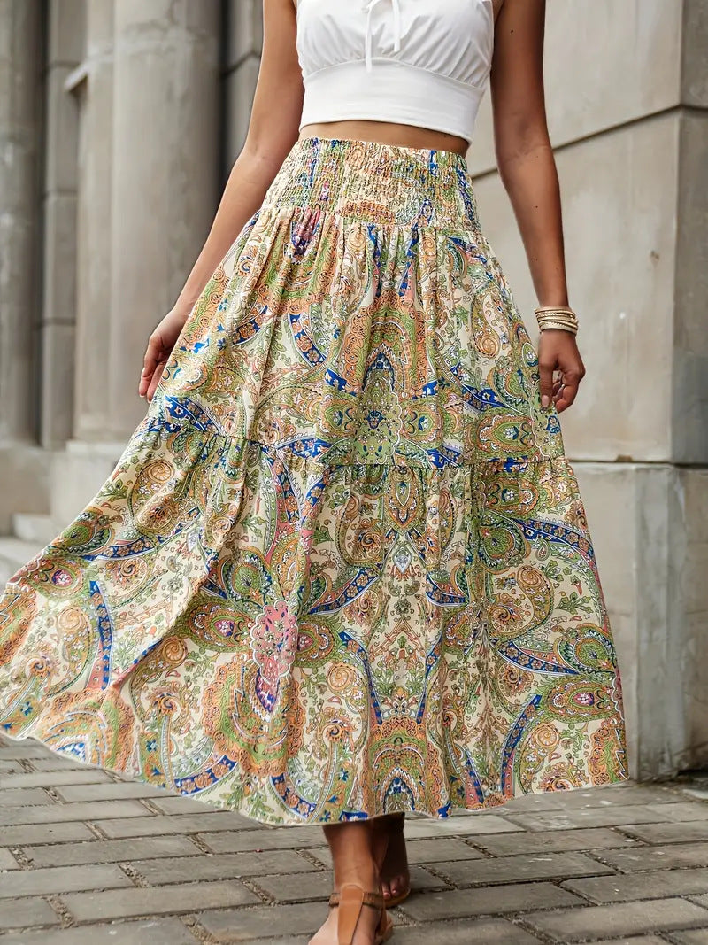 Frau in weißem Top und langem, buntem Boho-Rock mit Paisley-Muster, steht auf gepflastertem Weg vor grauen Säulen. Sommermode, Boho-Chic, Damenbekleidung.
