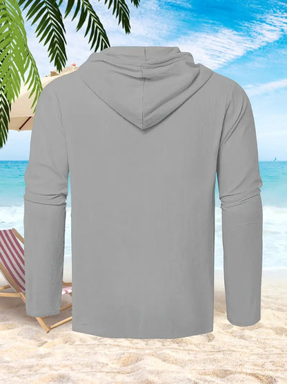 Graues Kapuzen-Sweatshirt am Strand, Palmen und Liegestuhl im Hintergrund, blauer Himmel und Meer. Ideal für Sommermode und Strandkleidung.