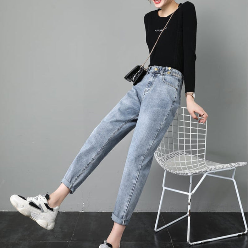 Frau in lässiger Mode: Schwarzes Langarmshirt, blaue Mom-Jeans, weiße Sneaker, schwarze Umhängetasche, sitzt auf weißem Drahtstuhl vor grauer Wand.