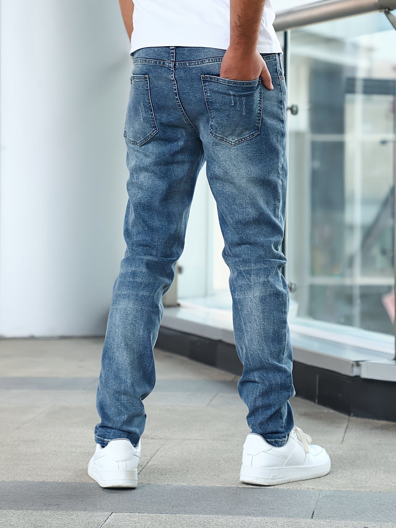 Mann in blauer Jeans und weißen Sneakern steht auf Balkon. Rückansicht, lässiger Stil, moderne Herrenmode, urbaner Hintergrund.