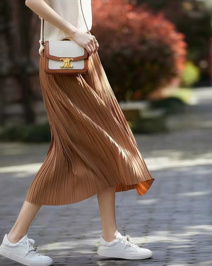 Frau mit braunem Plisseerock und weißem Top, trägt weiße Sneaker und braune Umhängetasche, geht auf gepflastertem Weg im Freien.