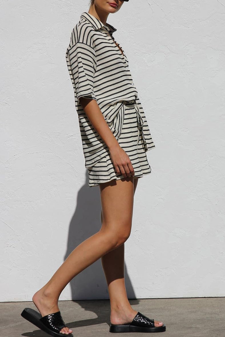 Frau in gestreiftem Sommeroutfit mit Shorts und Hemd, schwarze Sandalen, vor weißer Wand. Modetrend, lässiger Stil, Sommermode.