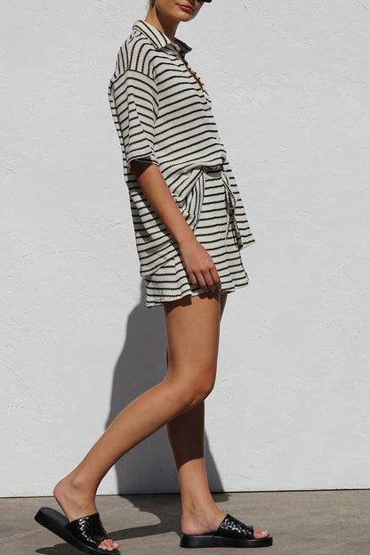 Frau in gestreiftem Sommeroutfit mit Shorts und Hemd, schwarze Sandalen, vor weißer Wand. Modetrend, lässiger Stil, Sommermode.