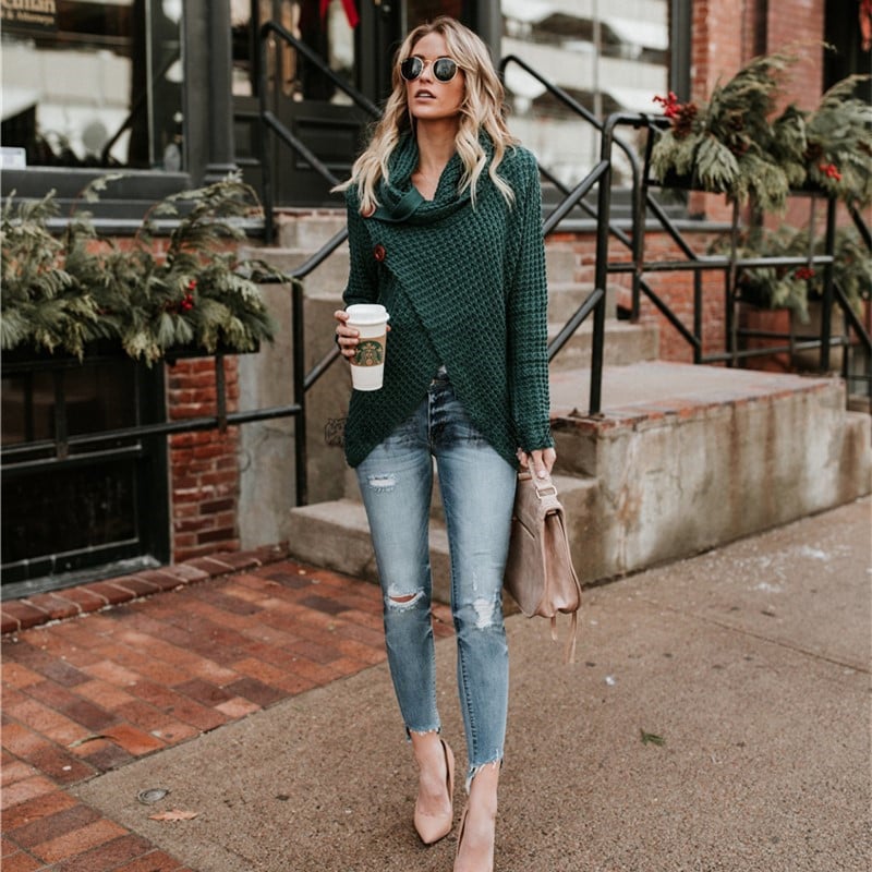 Frau in modischem, grünem Strickpullover und zerrissenen Jeans, hält Kaffeebecher, trägt Sonnenbrille, steht vor einem Café mit weihnachtlicher Dekoration.