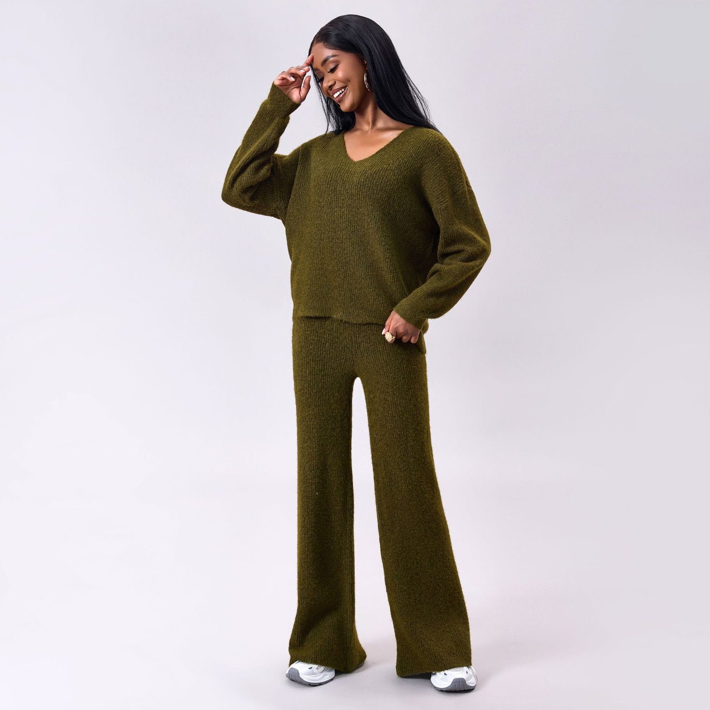 Frau in olivgrünem Strickpullover und Hose, lächelnd, mit langen schwarzen Haaren, trägt weiße Sneaker. Modetrend, bequeme Damenbekleidung.