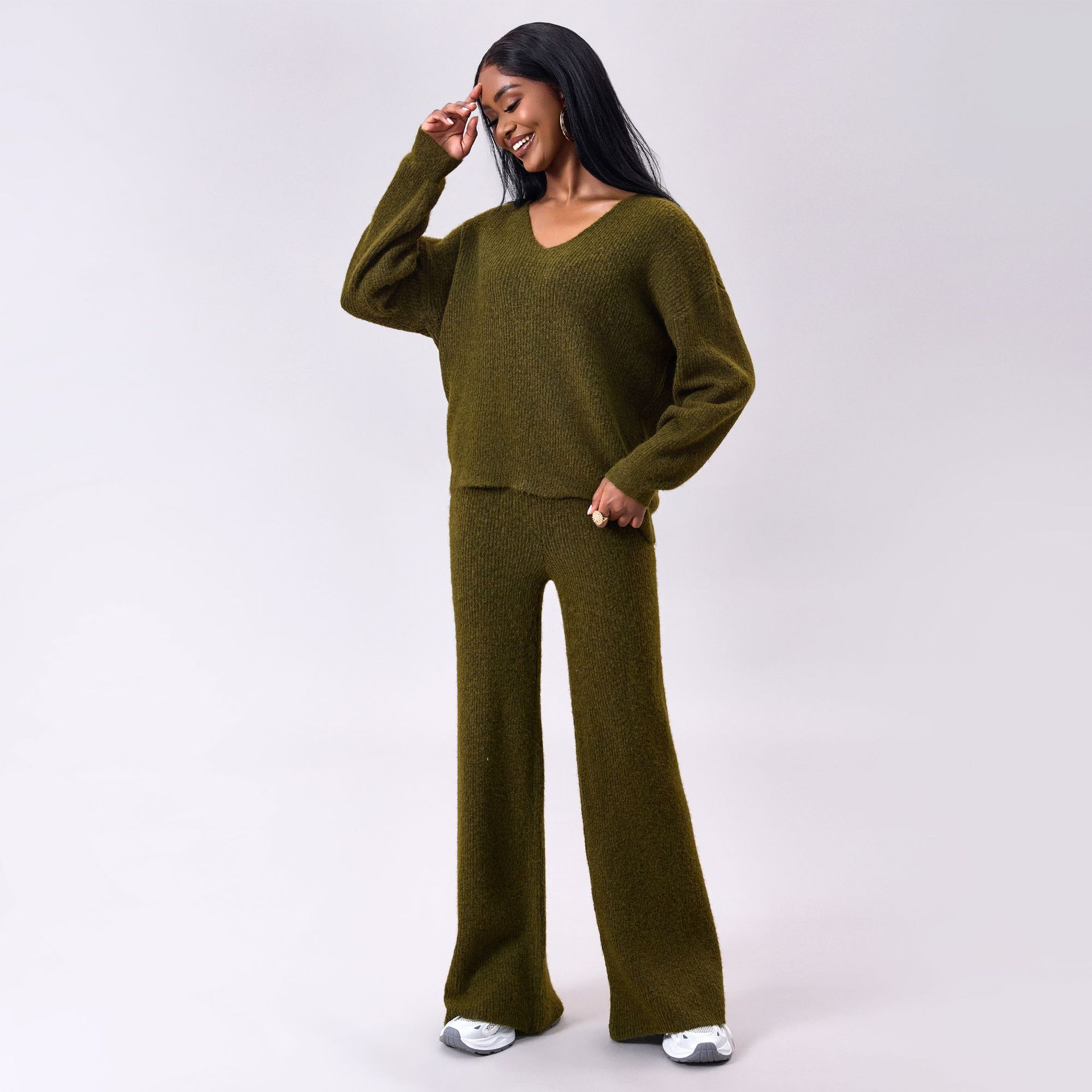 Frau in olivgrünem Strickpullover und Hose, lächelnd, mit langen schwarzen Haaren, trägt weiße Sneaker. Modetrend, bequeme Damenbekleidung.