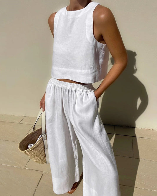 Frau in weißem Leinen-Zweiteiler mit ärmellosem Top und weiter Hose, hält Strohtasche. Sommermode, minimalistischer Stil, elegantes Outfit.
