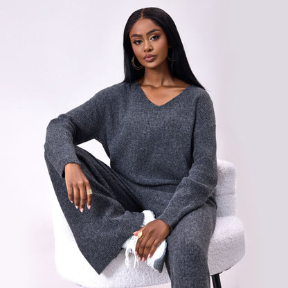 Frau in grauem Strickpullover und Hose sitzt auf einem weißen Stuhl, trägt goldene Ohrringe und weiße Sneaker, modisches Herbst-Outfit.