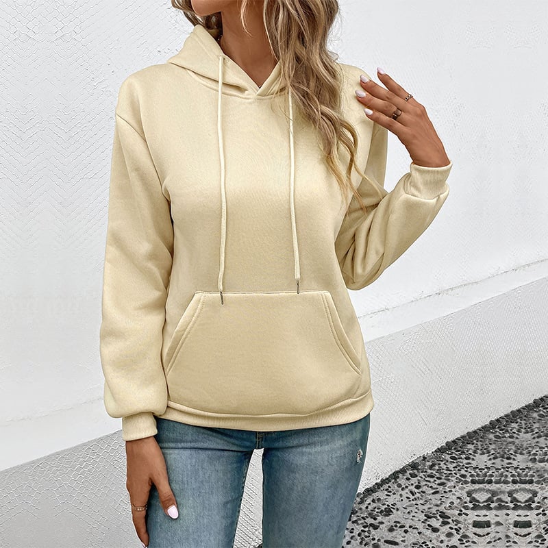 Frau in beigem Hoodie und blauer Jeans vor weißer Wand. Modetrend, lässiger Stil, Damenbekleidung, Kapuzenpullover, Freizeitlook.