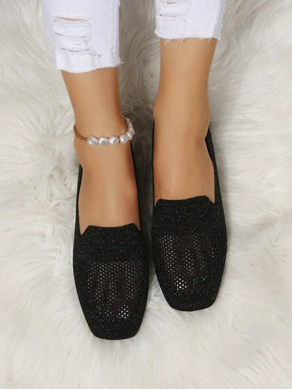 Schwarze glitzernde Damen-Loafer auf weißem Teppich, getragen mit zerrissenen weißen Jeans und Perlenfußkettchen. Elegante, modische Schuhe.