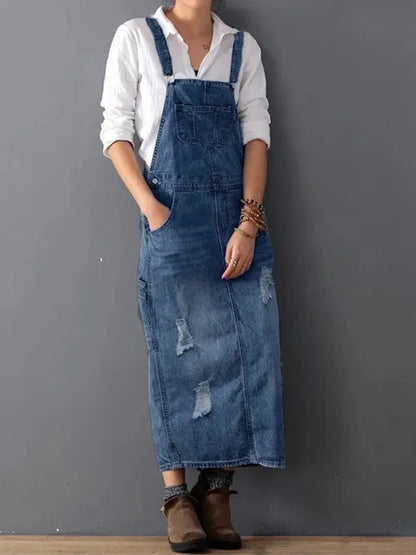 Frau in weißem Hemd und langer, zerrissener Jeans-Latzhose vor grauer Wand. Modetrend, lässiger Stil, Denim-Kleidung, Herbstmode.