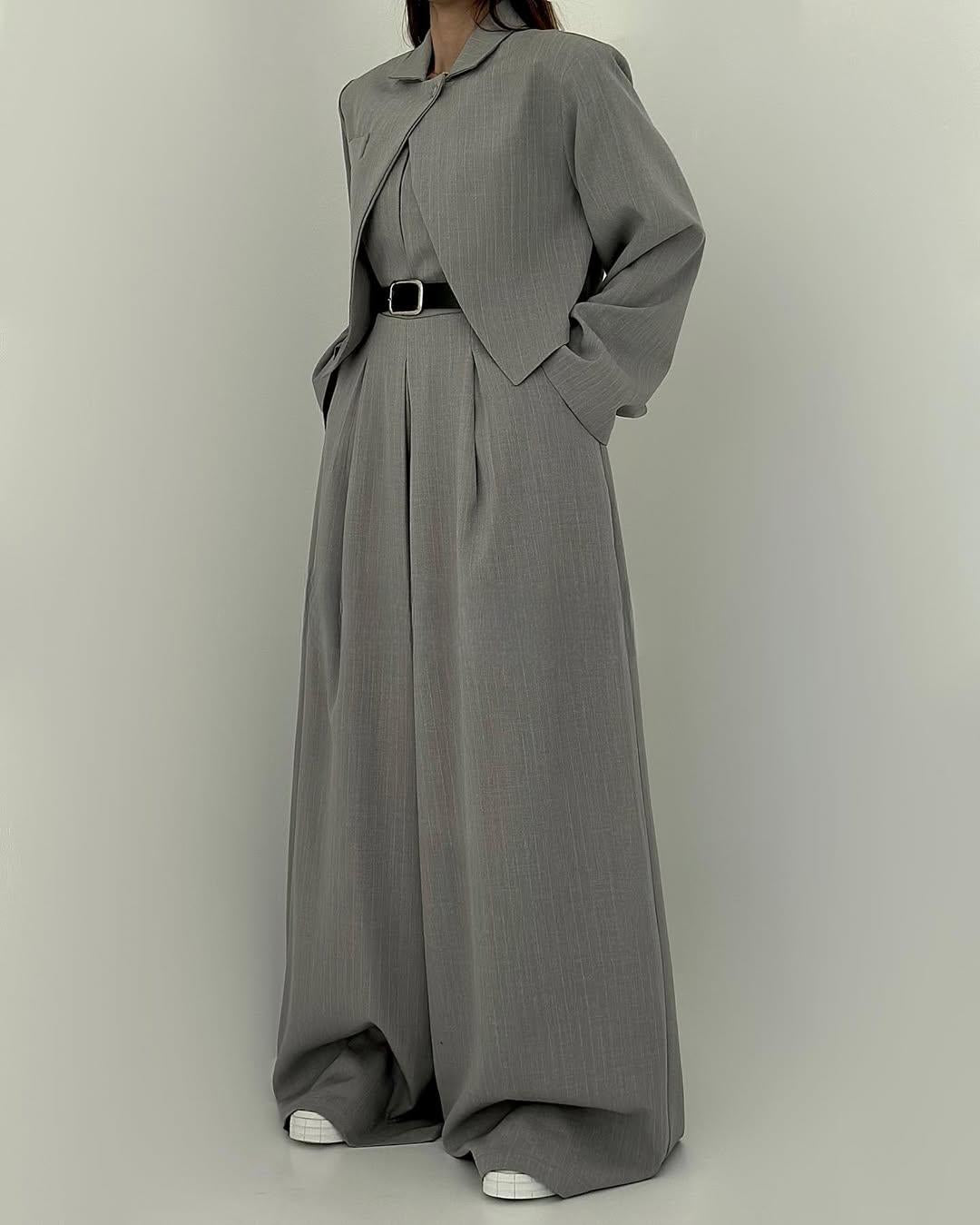 Grauer Damenanzug mit weitem Schnitt, bestehend aus Jacke und Hose, kombiniert mit schwarzem Gürtel und weißen Sneakers. Elegante Businessmode.