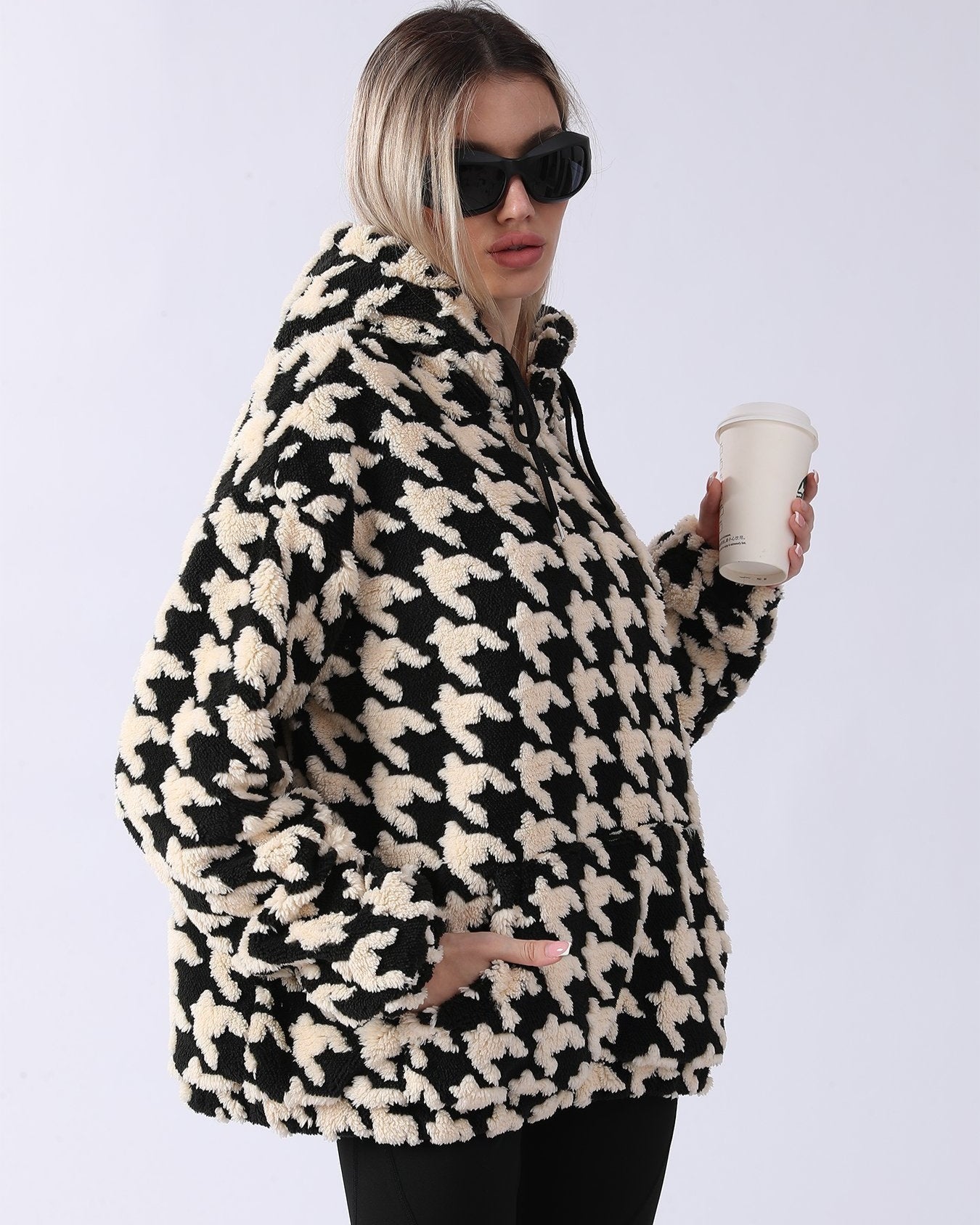 Frau in schwarz-weißem Hahnentritt-Fleece-Hoodie, hält Kaffeebecher, trägt Sonnenbrille. Modetrend, Wintermode, lässiger Stil.