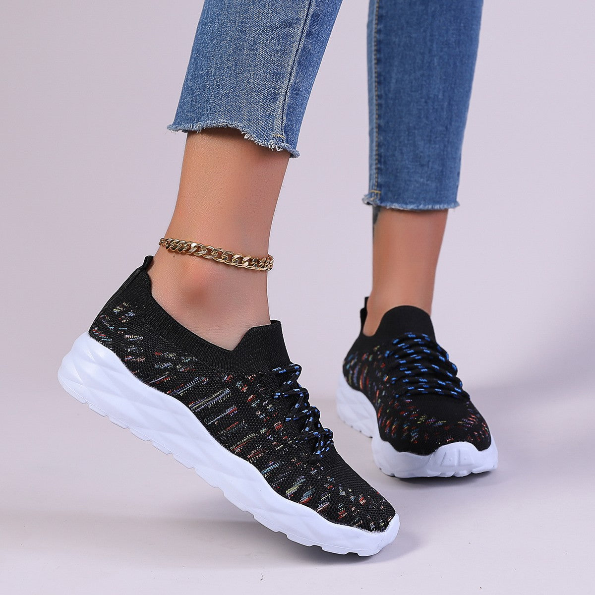 Schwarze Damen-Sneaker mit buntem Muster, weißer Sohle und blauen Schnürsenkeln, getragen mit blauer Jeans und goldener Fußkette.
