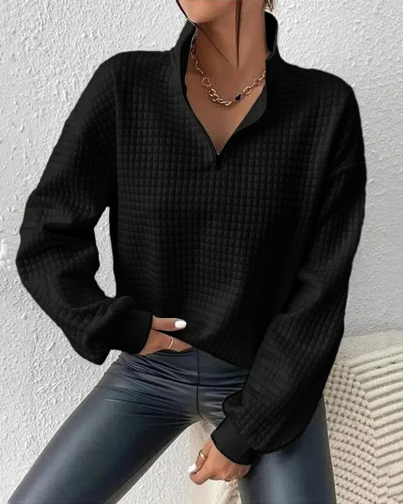 Frau in schwarzem, strukturiertem Pullover mit Reißverschluss und Kette, sitzt auf hellem Sofa. Modetrend, Damenmode, Herbstoutfit.
