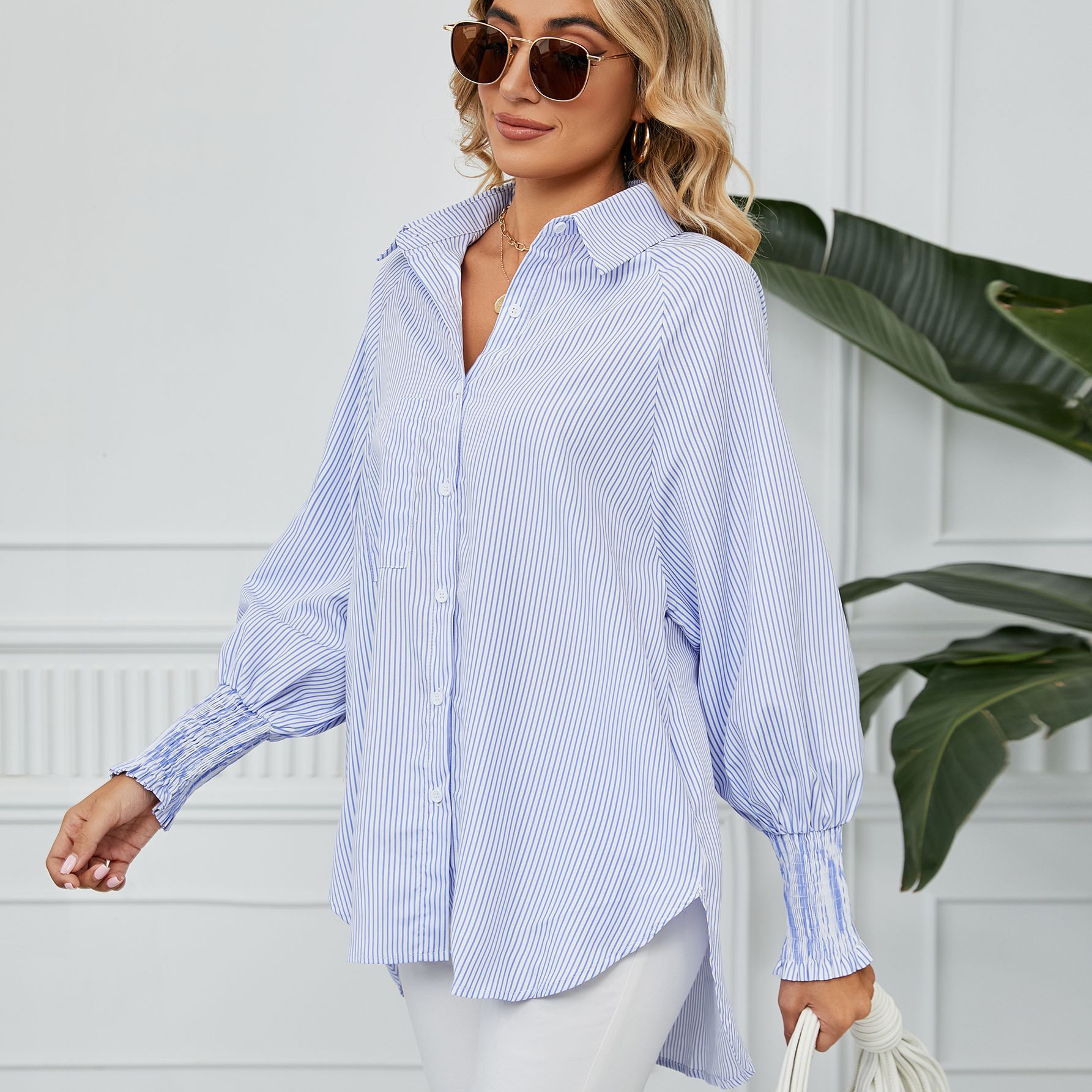 Frau in gestreifter Bluse und Sonnenbrille, lächelnd, vor weißer Wand mit Pflanzen. Modetrend, Damenmode, lässiger Stil, Sommeroutfit.