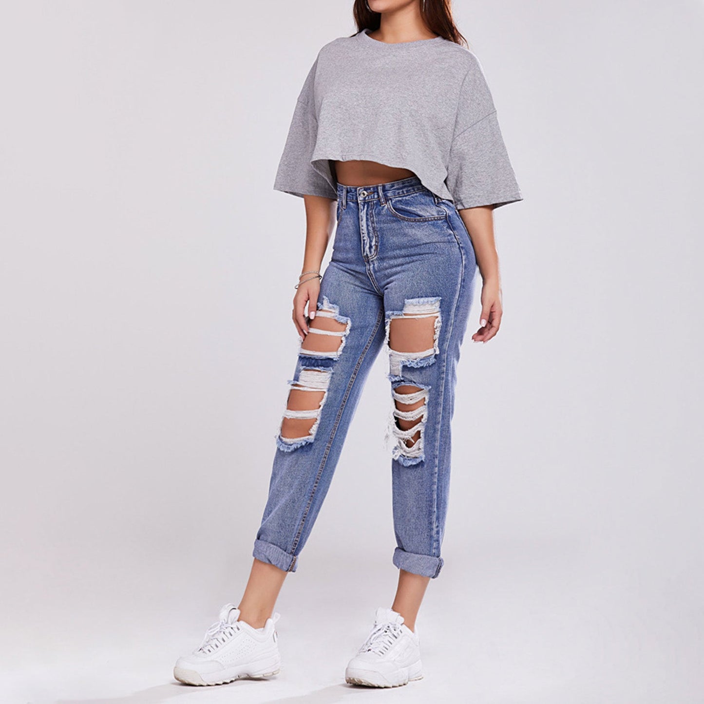 Frau in grauem Crop-Top und zerrissenen blauen Jeans, weiße Sneaker, modischer Streetwear-Look, lässiger Stil, trendige Damenmode.