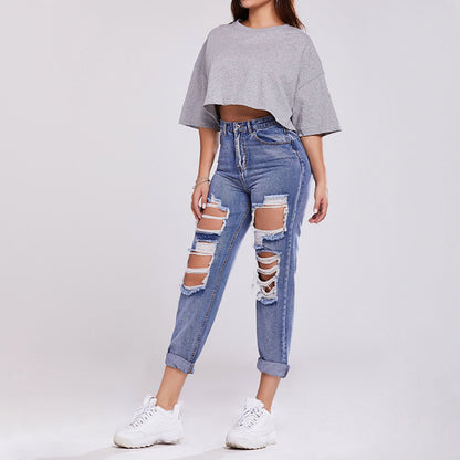 Frau in grauem Crop-Top und zerrissenen blauen Jeans, weiße Sneaker, modischer Streetwear-Look, lässiger Stil, trendige Damenmode.