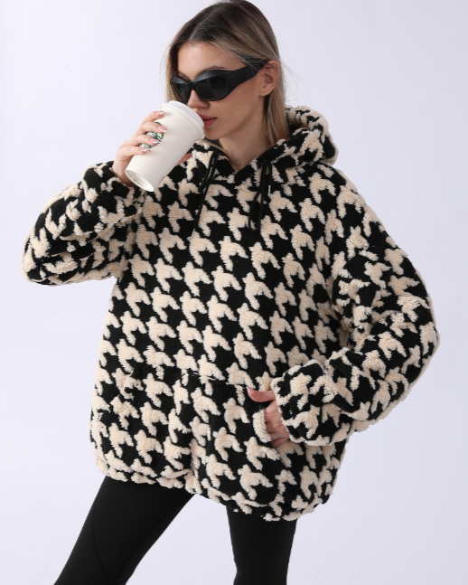 Frau in schwarz-weißem Hahnentritt-Hoodie trinkt Kaffee, trägt Sonnenbrille. Modetrend, Herbstmode, lässiger Stil, gemütlicher Look.