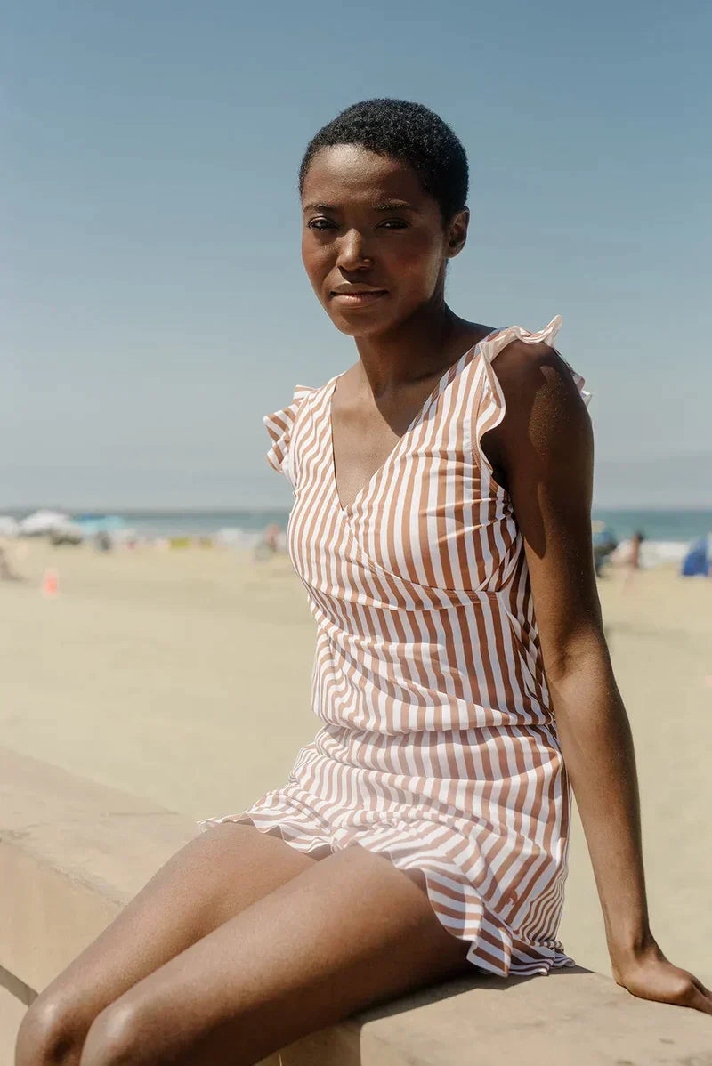 Eine Frau in einem gestreiften Sommerkleid sitzt am Strand. Klarer Himmel, Sand und Meer im Hintergrund. Sommermode, Strandtag, Freizeitkleidung.