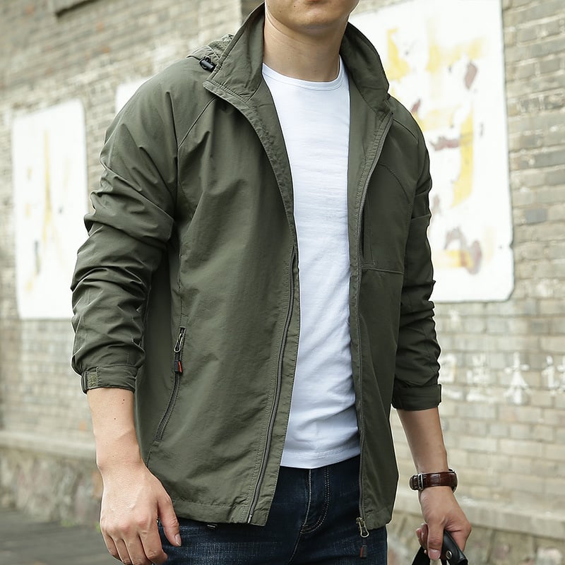 Mann in olivgrüner Windjacke und weißem T-Shirt vor Backsteinwand. Modische Herrenbekleidung, lässiger Stil, Outdoor-Jacke.