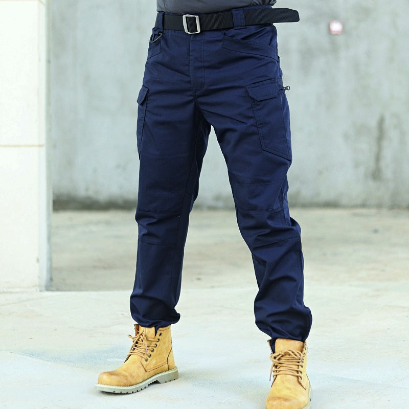 Mann trägt dunkelblaue taktische Hose und gelbe Arbeitsstiefel, steht auf Betonboden. Outdoor-Mode, robuste Kleidung, Herrenbekleidung.