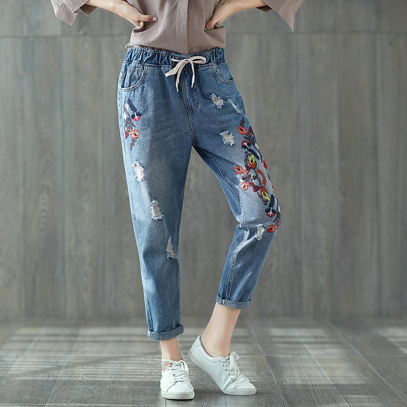 Damenjeans mit Blumenstickerei, lässiger Stil, blaue Denim-Hose, modische Risse, elastischer Bund, weiße Sneaker, Holzfußboden.