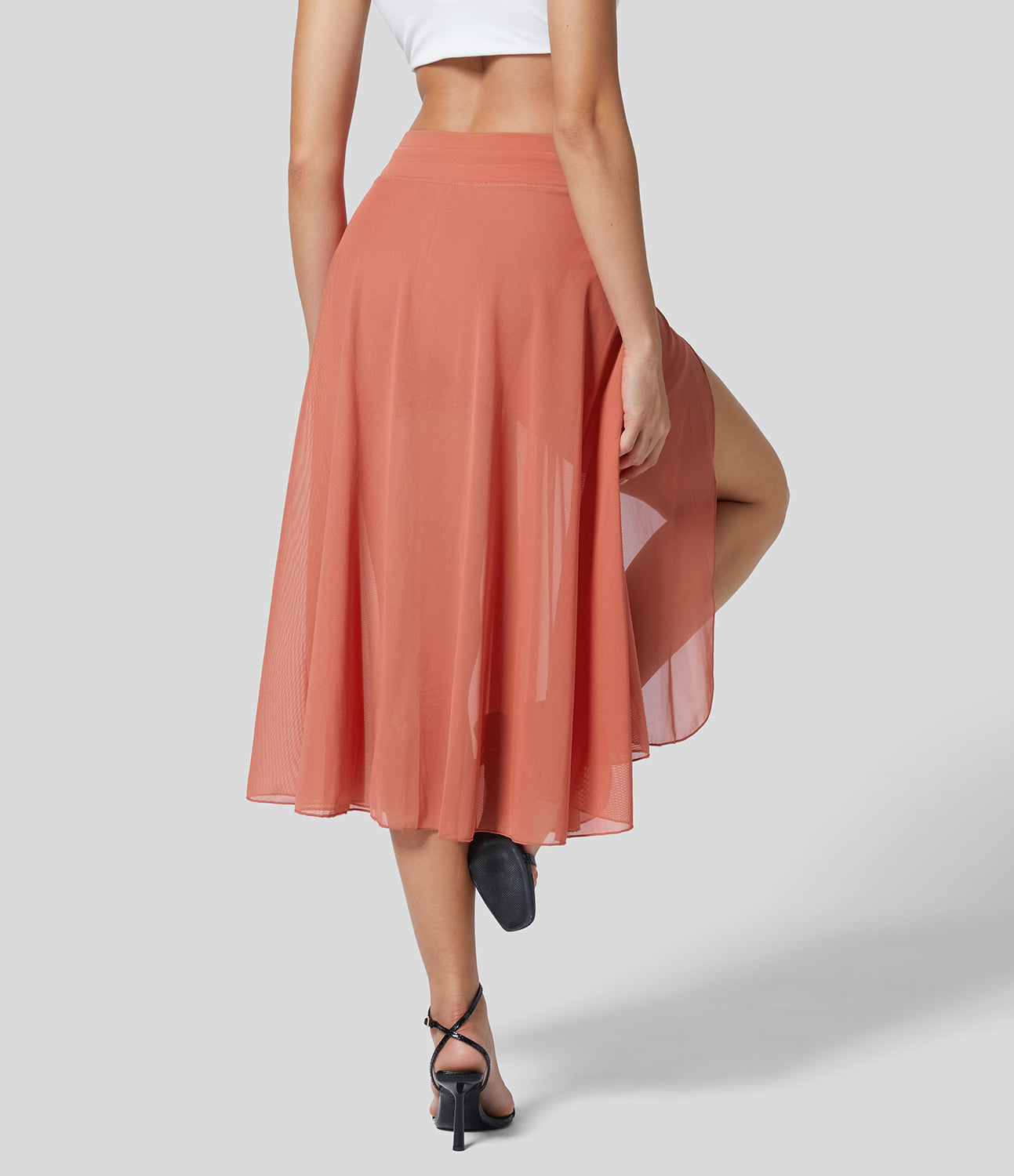 Frau in rotem, fließendem Midirock und weißem Top, zeigt Bein. Elegante Sandalen, grauer Hintergrund. Modetrend, Sommeroutfit, Damenmode.