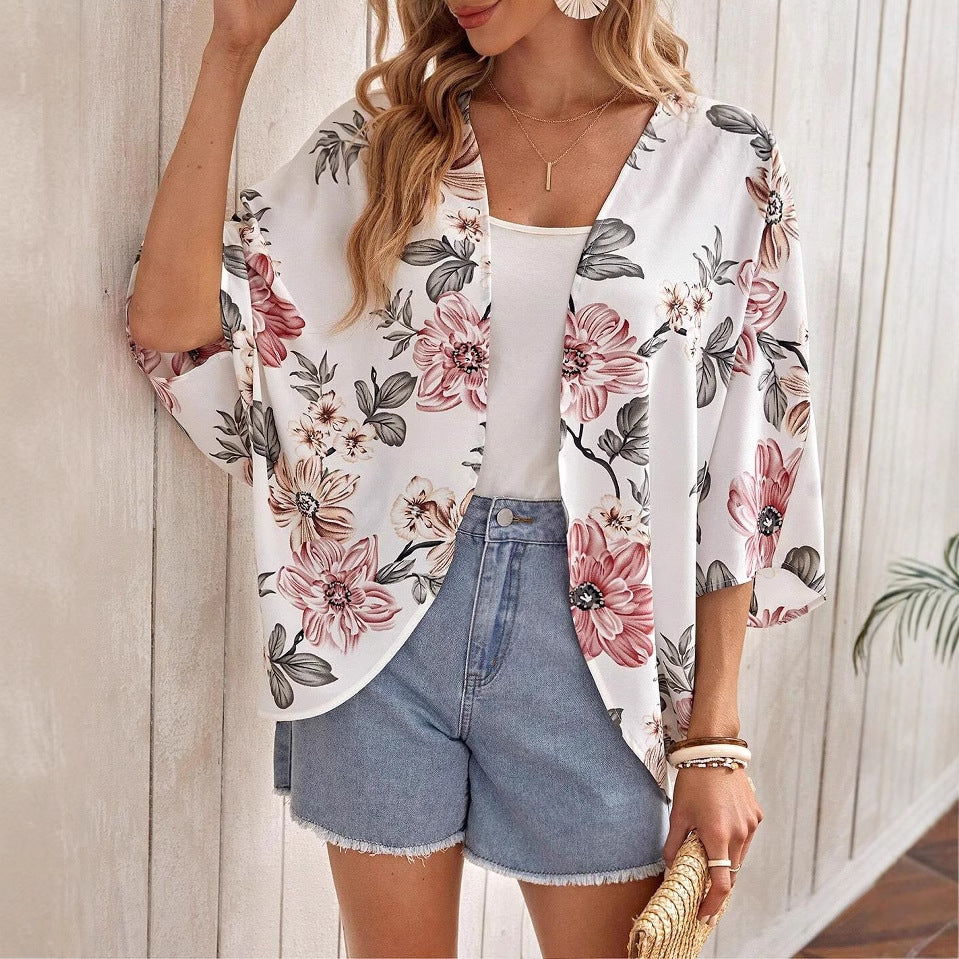 Frau in floralem Kimono, weißem Top und Jeansshorts, hält Strohtasche. Sommermode, lässiger Stil, Blumenmuster, trendige Damenbekleidung.