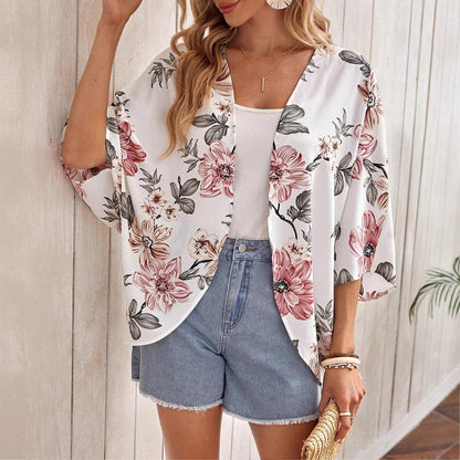 Frau in floralem Kimono, weißem Top und Jeansshorts, hält Strohtasche. Sommermode, lässiger Stil, Blumenmuster, trendige Damenbekleidung.