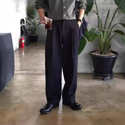 Mann in schwarzer Hose und Hemd hält Getränk, steht in modernem Raum mit Pflanzen. Stilvolle Herrenmode, lässiger Look, Indoor-Umgebung.