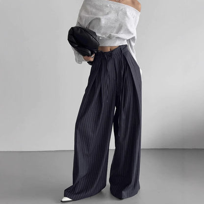 Frau in grauem Off-Shoulder-Pullover und gestreifter, weiter Hose, hält schwarze Tasche. Modetrend, lässiger Stil, minimalistische Kleidung.