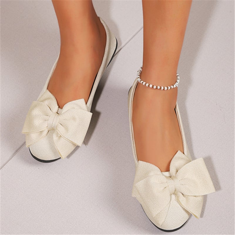 Weiße Ballerinas mit großer Schleife und Perlenfußkettchen auf hellem Boden. Modische Damenschuhe, elegantes Design, trendige Accessoires.