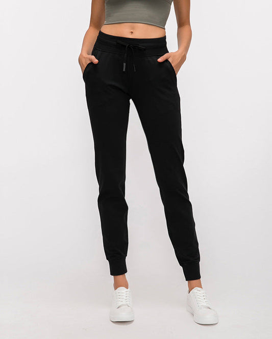 Frau in schwarzer Jogginghose und weißen Sneakers, graues Top, Hände in den Taschen. Bequeme Sportbekleidung, lässiger Stil.