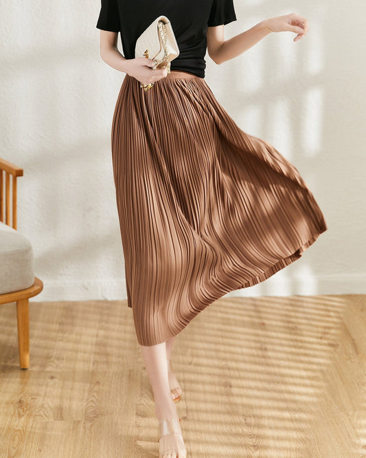 Frau in schwarzem Oberteil und braunem Plisseerock, hält eine weiße Clutch. Elegante Mode, Sommeroutfit, Innenraum, Holzboden.