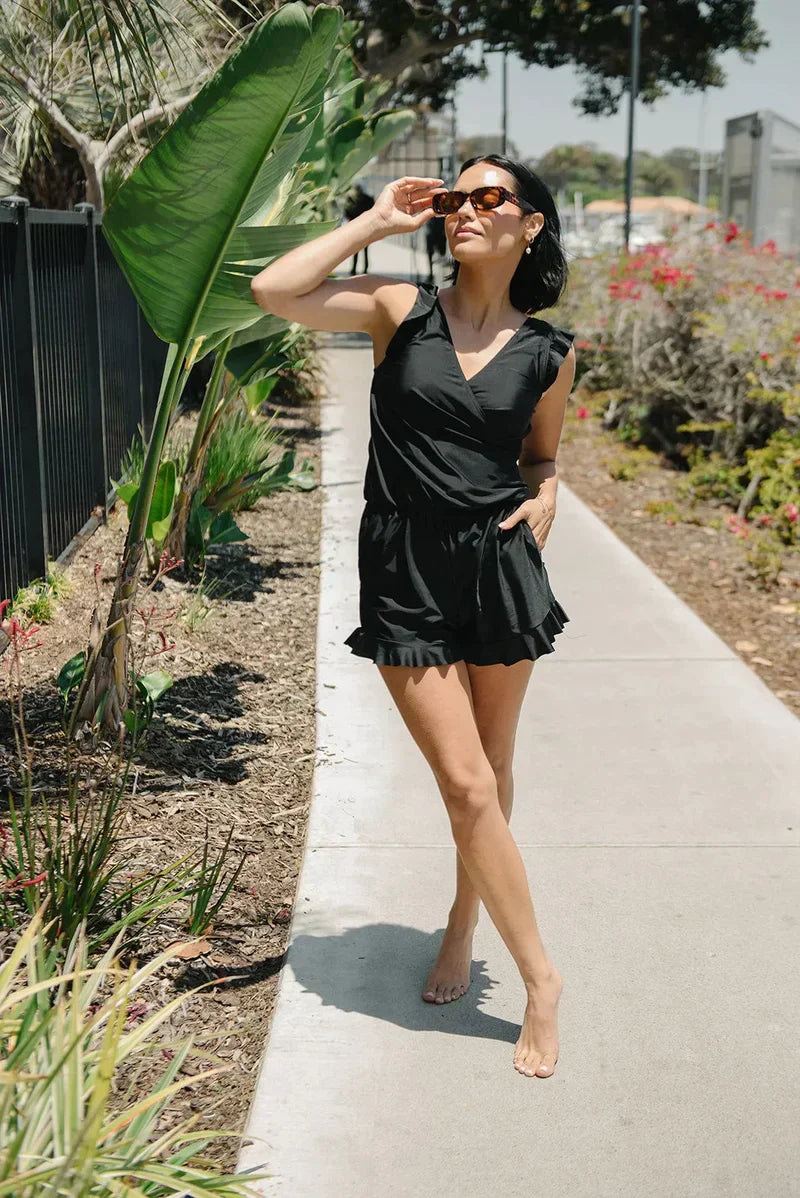 Frau in schwarzem Sommerkleid und Sonnenbrille spaziert barfuß auf einem Gehweg, umgeben von Pflanzen und sonnigem Wetter.