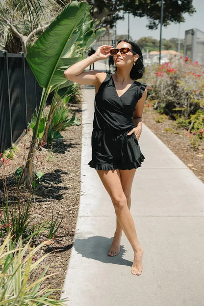 Frau in schwarzem Sommerkleid und Sonnenbrille spaziert barfuß auf einem Gehweg, umgeben von Pflanzen und sonnigem Wetter.