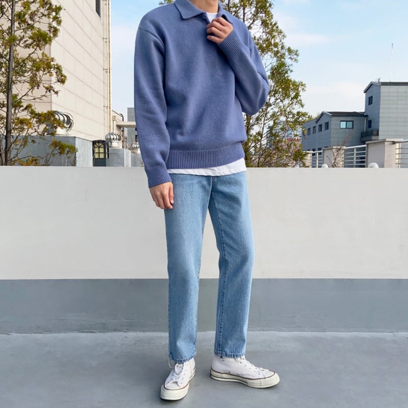 Mann in blauem Pullover und Jeans steht auf Dachterrasse, trägt weiße Sneakers. Urbaner Hintergrund mit Gebäuden und Bäumen.