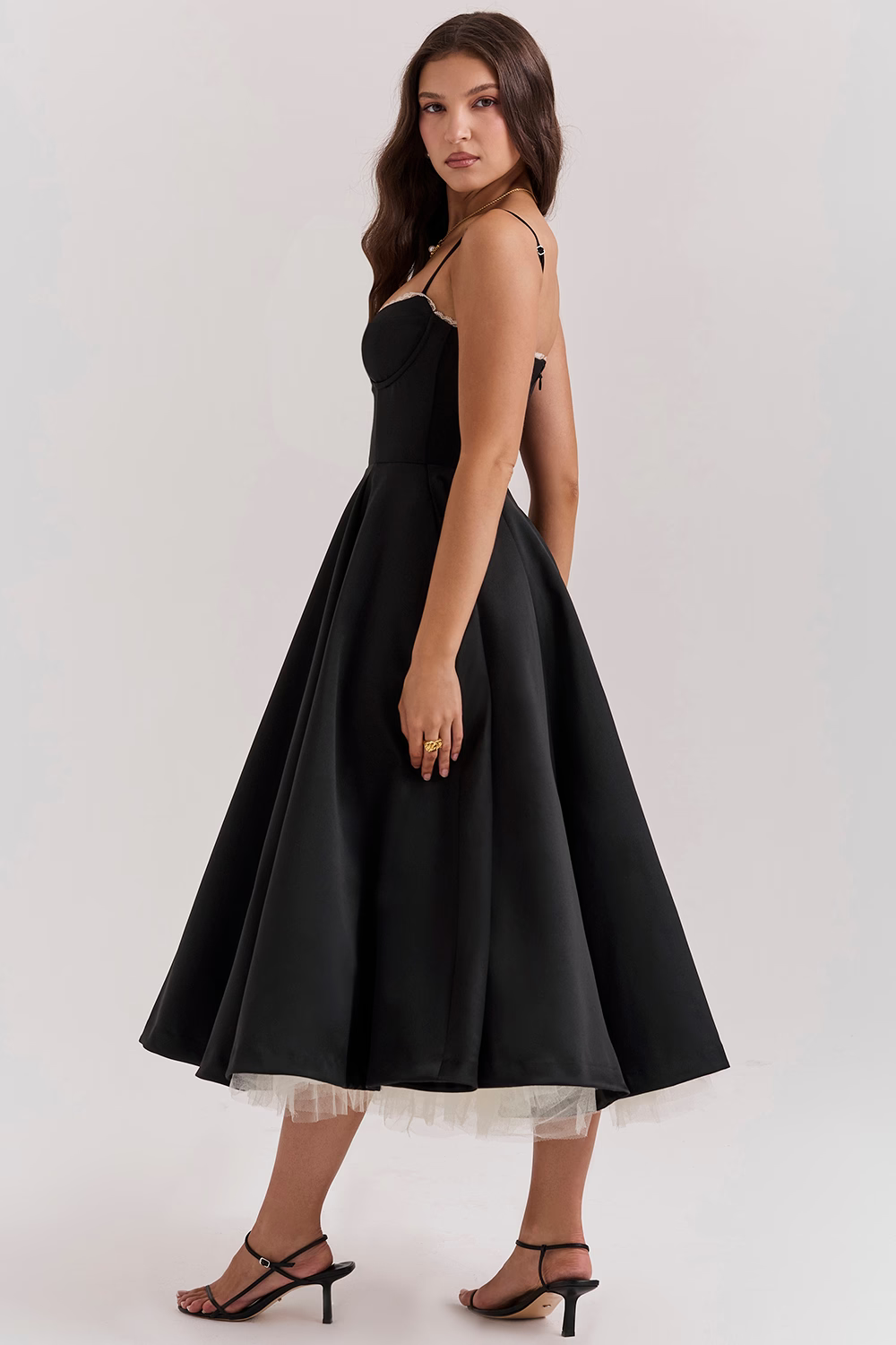 Frau in elegantem, schwarzem Abendkleid mit Spaghettiträgern und ausgestelltem Rock, seitlich stehend, auf weißem Hintergrund.