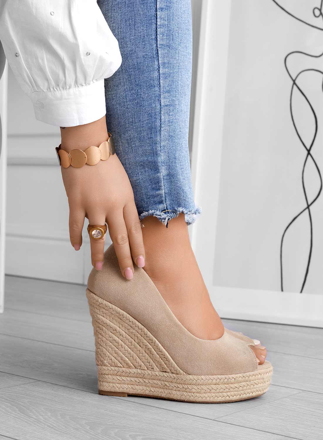 Frau in blauer Jeans und weißer Bluse trägt beige Keilabsatz-Schuhe, Schmuck am Handgelenk, modernes Design im Hintergrund.