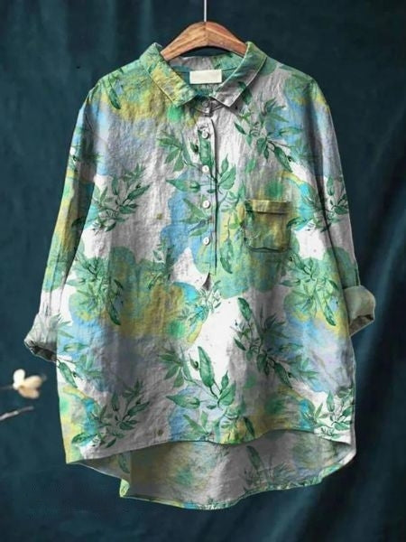 Grüne Damenbluse mit floralem Muster, Knopfleiste und Brusttasche, aus Leinenstoff, auf Holzkleiderbügel vor dunkelblauem Hintergrund.