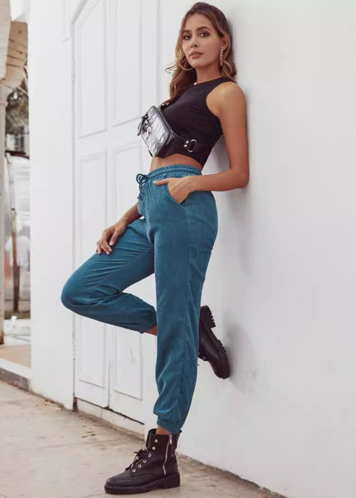 Frau in schwarzem Top und blauen Jogginghosen lehnt an weißer Wand, trägt schwarze Stiefel und Bauchtasche. Streetwear, lässiger Look, Mode.