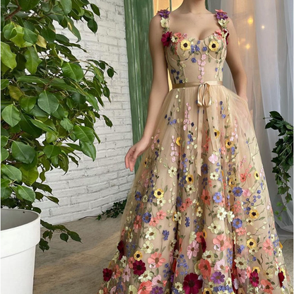 Frau in elegantem, ärmellosem Kleid mit bunten Blumenstickereien, steht neben grünen Pflanzen vor einer weißen Backsteinwand.
