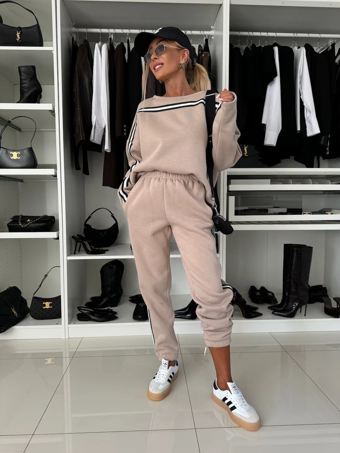 Frau in beige Jogginganzug und weißen Sneakers posiert vor einem Kleiderschrank mit schwarzen Stiefeln und Kleidung. Sportlich, modisch, lässig.