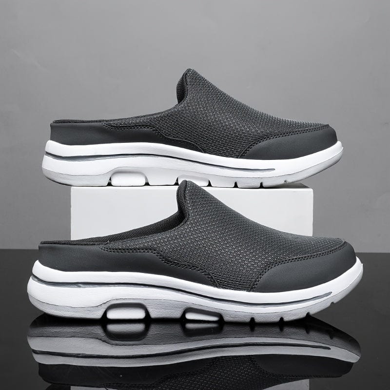 Graue Herren-Sneaker-Slipper mit weißer Sohle, atmungsaktivem Mesh-Obermaterial, auf weißem Podest vor grauem Hintergrund. Bequeme Freizeitschuhe.