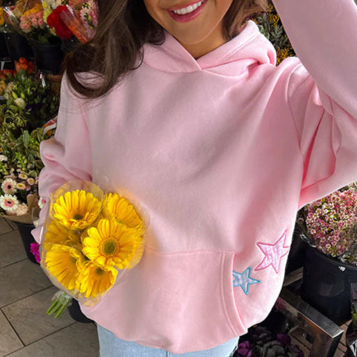 Frau in rosa Hoodie hält gelben Blumenstrauß, umgeben von Blumenladen. Frühlingsmode, lässiger Stil, frische Blumen.